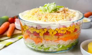 Omas Schichtsalat in einer Glasbowle, geschichtet mit Lauch, Karotten, Ananas, Tomaten, Mais und Schinken, gekrönt mit Mayonnaise, Käse und Frühlingszwiebeln.