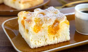 Ein Stück saftiger Buttermilchkuchen mit Mandarinen, bestäubt mit Puderzucker, auf einem braunen Teller neben einer Tasse Kaffee.