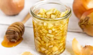 Zwiebel-Honig-Saft in einem Glas, daneben Zwiebeln und ein Honiglöffel. Ein natürliches Hausmittel.
