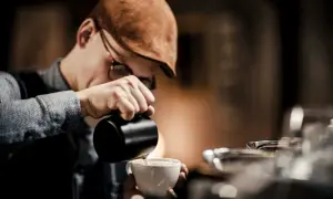 Barista mit Schiebermütze gießt Milch in eine Tasse, um perfekten Kaffee-Schaum zu kreieren.