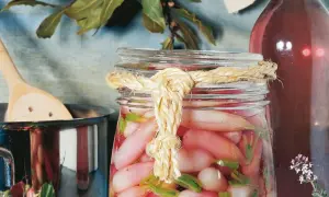 Ein Glas mit gepickelten Radieschen, verziert mit einem Seil. Daneben ein Topf mit einem Holzlöffel und eine Flasche mit roter Flüssigkeit.