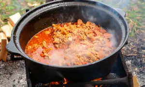 Herzhafte Eintopf-Rezepte im Kessel über offenem Feuer. Dampf steigt auf, während Fleisch und Gemüse in der roten Sauce köcheln.