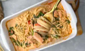 Eine Gabel hebt eine Portion cremige Pasta mit Lachs und Tomaten aus einer Auflaufform.
