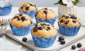 Fünf saftige, vegane Blaubeermuffins in blauen Papierförmchen auf einem weißen Teller. Frische Blaubeeren und eine kleine Blumendekoration runden das Bild ab.