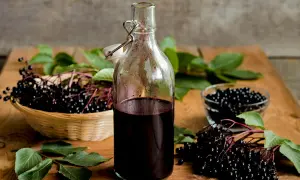 Flasche mit dunklem Holundersaft, daneben ein Korb mit frischen Holunderbeeren. Perfekt für ein Holundersaft Rezept.