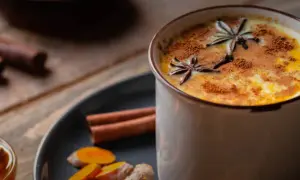 Eine Tasse goldene Milch, garniert mit Sternanis und Zimt. Daneben Zutaten für das goldene Milch Rezept: Kurkuma, Ingwer, Zimtstangen und Honig.
