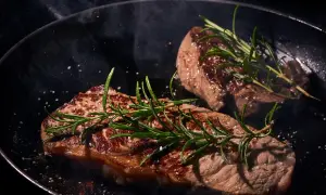 Zwei saftige Steaks braten in einer Pfanne mit Rosmarin und grobem Salz. Dampf steigt auf und verströmt ein köstliches Aroma.