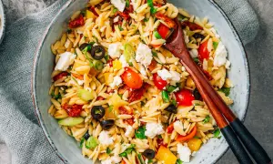 Ein bunter Kritharaki Salat mit Tomaten, Oliven, Feta und Paprika, serviert in einer Schüssel mit Holzstäbchen.