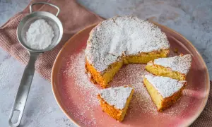 Mallorquinischer Mandelkuchen, originalrezept: Ein saftiger Kuchen mit Puderzucker bestäubt, auf einem rosa Teller, daneben ein Sieb mit Puderzucker.