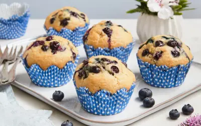 Fünf saftige, vegane Blaubeermuffins in blauen Papierförmchen auf einem weißen Teller. Frische Blaubeeren und eine kleine Blumendekoration runden das Bild ab.