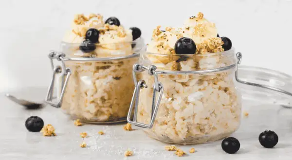 Zwei Gläser mit Overnight Oats, garniert mit Bananen, Blaubeeren und Granola. Perfekt für ein schnelles Frühstück.