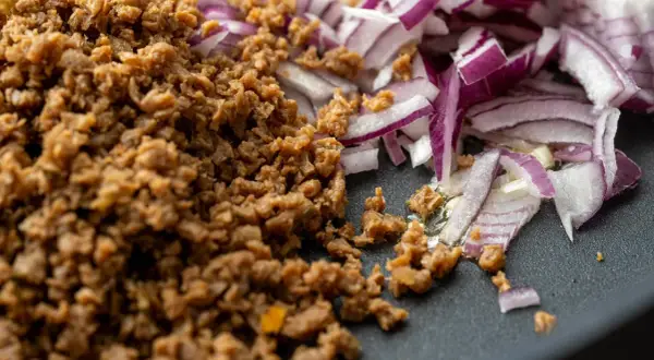 Gebratenes Hackfleischersatz-Granulat neben roten Zwiebeln in einer Pfanne.