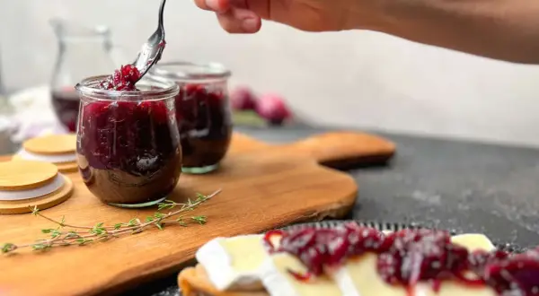 Eine Hand schöpft mit einem Löffel köstliche zwiebelmarmelade einfach aus einem Glas. Dazu passt Brot mit Käse.