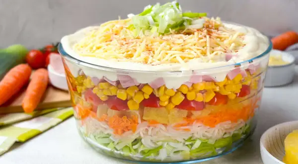 Omas Schichtsalat in einer Glasbowle, geschichtet mit Lauch, Karotten, Ananas, Tomaten, Mais und Schinken, gekrönt mit Mayonnaise, Käse und Frühlingszwiebeln.