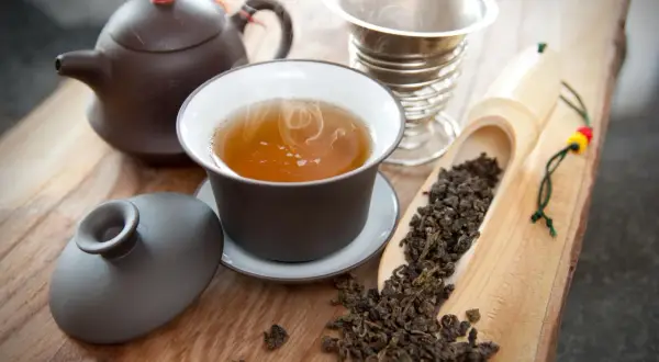 Dampfender Oolong Tee in einer Tasse, daneben eine Teekanne, ein Sieb und ein Löffel mit losem Tee. Die Oolong Tee Wirkung kann belebend sein.