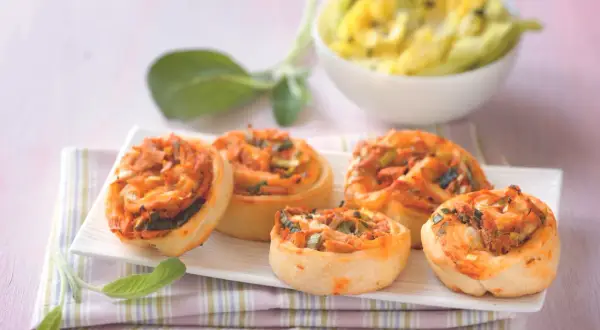 Fünf leckere Pizzaschnecken Thunfisch mit Tomaten und Lauchzwiebeln auf einem weißen Teller, daneben ein Schälchen mit gelbem Dip.
