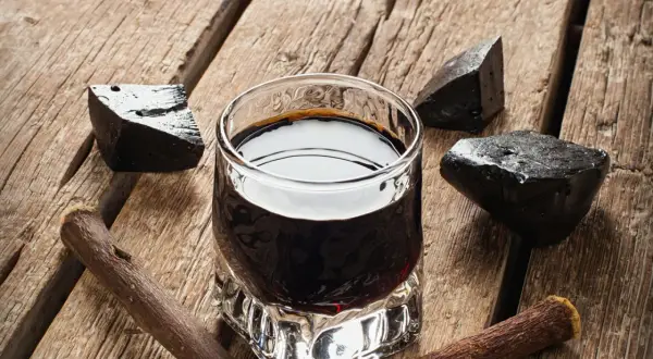 Ein Glas dunkler Flüssigkeit, umgeben von Lakritzstücken und -wurzeln. Perfekt für alle, die **Lakritz Schnaps selber machen** möchten.