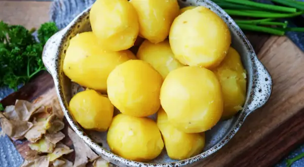 Schüssel mit frisch gekochten Kartoffeln, die mit Schale serviert werden. Daneben liegen Kartoffelschalen und frische Kräuter.
