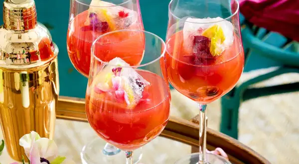 Erfrischendes Aperol Spritz Rezept: Drei Gläser mit leuchtend rotem Aperol, Eiswürfeln und Fruchtgarnitur. Ein goldener Shaker steht bereit.
