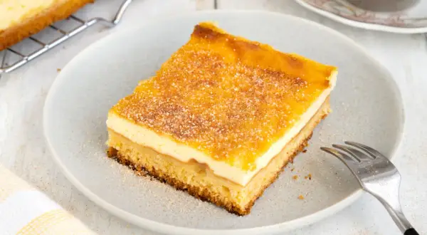 Ein Stück von Omas Schmandkuchen vom Blech, bestreut mit Zucker und Zimt, liegt auf einem Teller.
