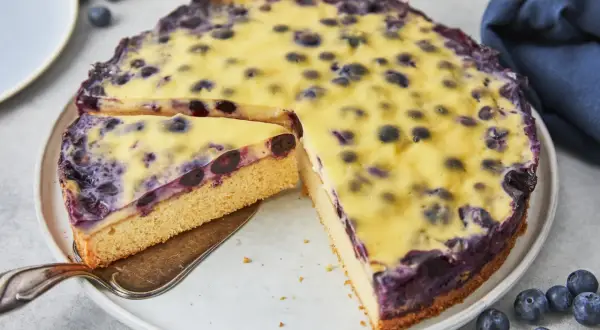 Ein saftiger Heidelbeerkuchen mit cremigem Vanillepudding, ein Stück wird gerade herausgehoben.