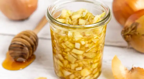 Zwiebel-Honig-Saft in einem Glas, daneben Zwiebeln und ein Honiglöffel. Ein natürliches Hausmittel.