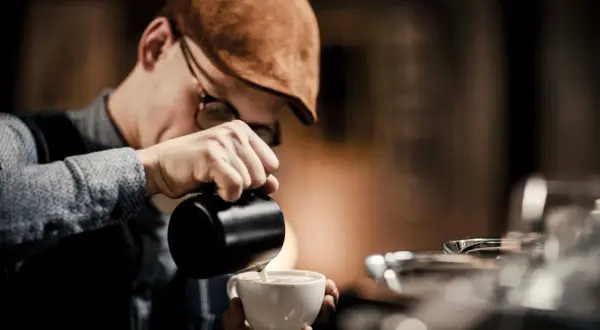 Barista mit Schiebermütze gießt Milch in eine Tasse, um perfekten Kaffee-Schaum zu kreieren.