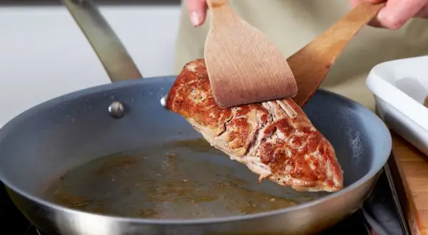 Ein saftiges Schweinefilet wird mit zwei Holzspateln in einer Pfanne gewendet, um es perfekt zu braten.
