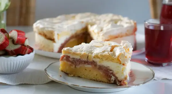 Ein saftiger Rhabarberkuchen mit Baiser, wie ihn Landfrauen backen. Ein Stück auf einem Teller, daneben frischer Rhabarber.