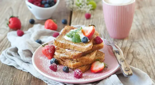 Ein Stapel goldbrauner French Toast, bestäubt mit Puderzucker, garniert mit Beeren und Sahne. Ein köstlicher Start in den Tag!