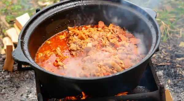 Herzhafte Eintopf-Rezepte im Kessel über offenem Feuer. Dampf steigt auf, während Fleisch und Gemüse in der roten Sauce köcheln.
