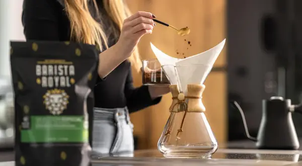 Frau gibt mit Löffel Kaffeepulver in den Filter, um Filterkaffee zu kochen. Daneben steht eine Packung Barista Royal Kaffee.