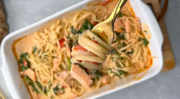 Eine Gabel hebt eine Portion cremige Pasta mit Lachs und Tomaten aus einer Auflaufform.