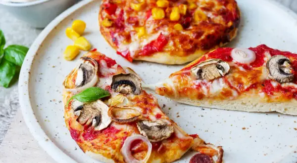 Zwei kleine Pizzen, eine mit Pilzen und Zwiebeln, die andere mit Mais und Paprika, warten darauf, aufgebacken zu werden.