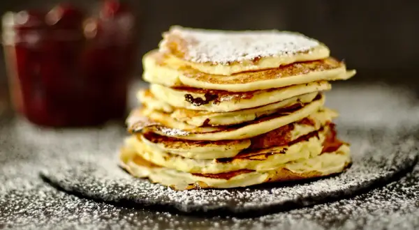 Ein Stapel fluffiger Quark Pfannkuchen, bestäubt mit Puderzucker, neben einem Glas Marmelade.