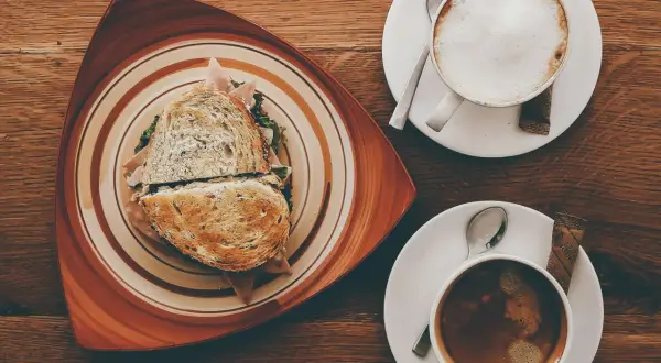 Ein Sandwich und zwei Kaffees auf einem Holztisch. Der Kaffee könnte mit Kaffeesirup verfeinert werden.