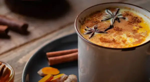 Eine Tasse goldene Milch, garniert mit Sternanis und Zimt. Daneben Zutaten für das goldene Milch Rezept: Kurkuma, Ingwer, Zimtstangen und Honig.