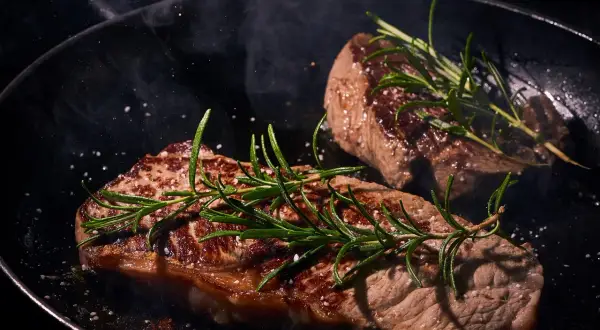 Zwei saftige Steaks braten in einer Pfanne mit Rosmarin und grobem Salz. Dampf steigt auf und verströmt ein köstliches Aroma.