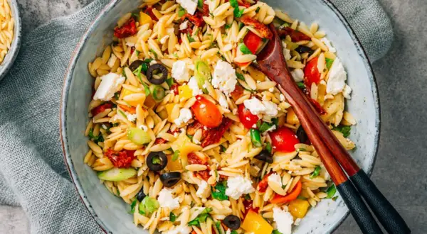 Ein bunter Kritharaki Salat mit Tomaten, Oliven, Feta und Paprika, serviert in einer Schüssel mit Holzstäbchen.