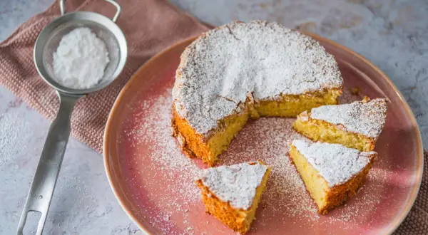 Mallorquinischer Mandelkuchen, originalrezept: Ein saftiger Kuchen mit Puderzucker bestäubt, auf einem rosa Teller, daneben ein Sieb mit Puderzucker.