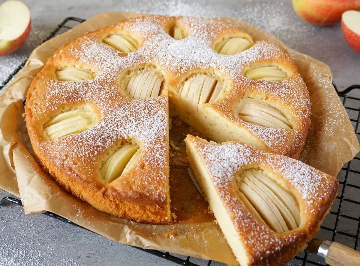 Ein goldbrauner Apfelkuchen, bestäubt mit Puderzucker, mit sichtbaren Apfelscheiben. Ein Stück ist herausgeschnitten. Ein gesunder Genuss!