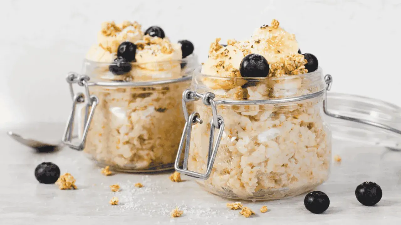 Zwei Gläser mit Overnight Oats, garniert mit Bananen, Blaubeeren und Granola. Perfekt für ein schnelles Frühstück.