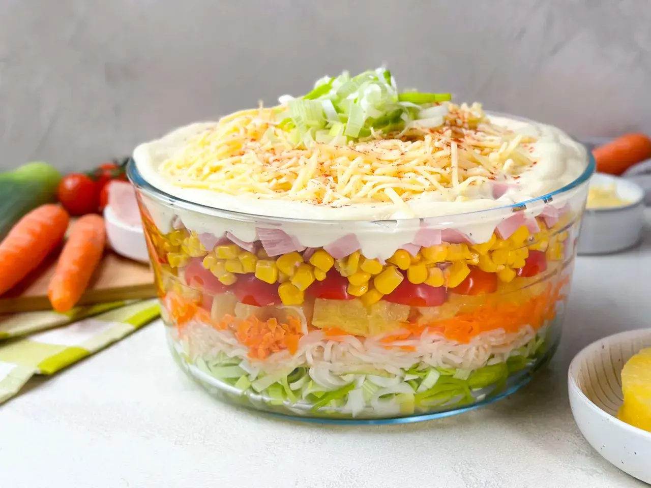 Omas Schichtsalat in einer Glasbowle, geschichtet mit Lauch, Karotten, Ananas, Tomaten, Mais und Schinken, gekrönt mit Mayonnaise, Käse und Frühlingszwiebeln.