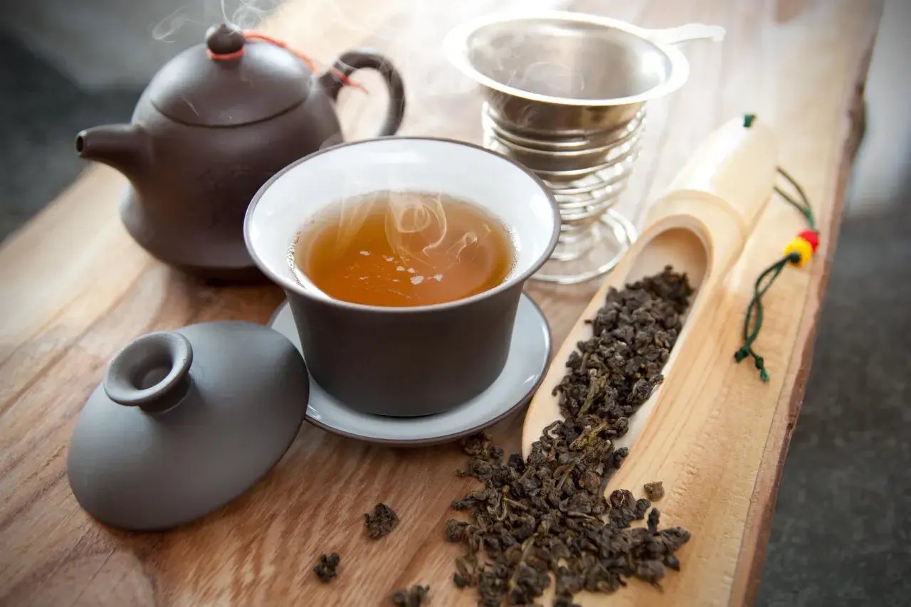 Dampfender Oolong Tee in einer Tasse, daneben eine Teekanne, ein Sieb und ein Löffel mit losem Tee. Die Oolong Tee Wirkung kann belebend sein.