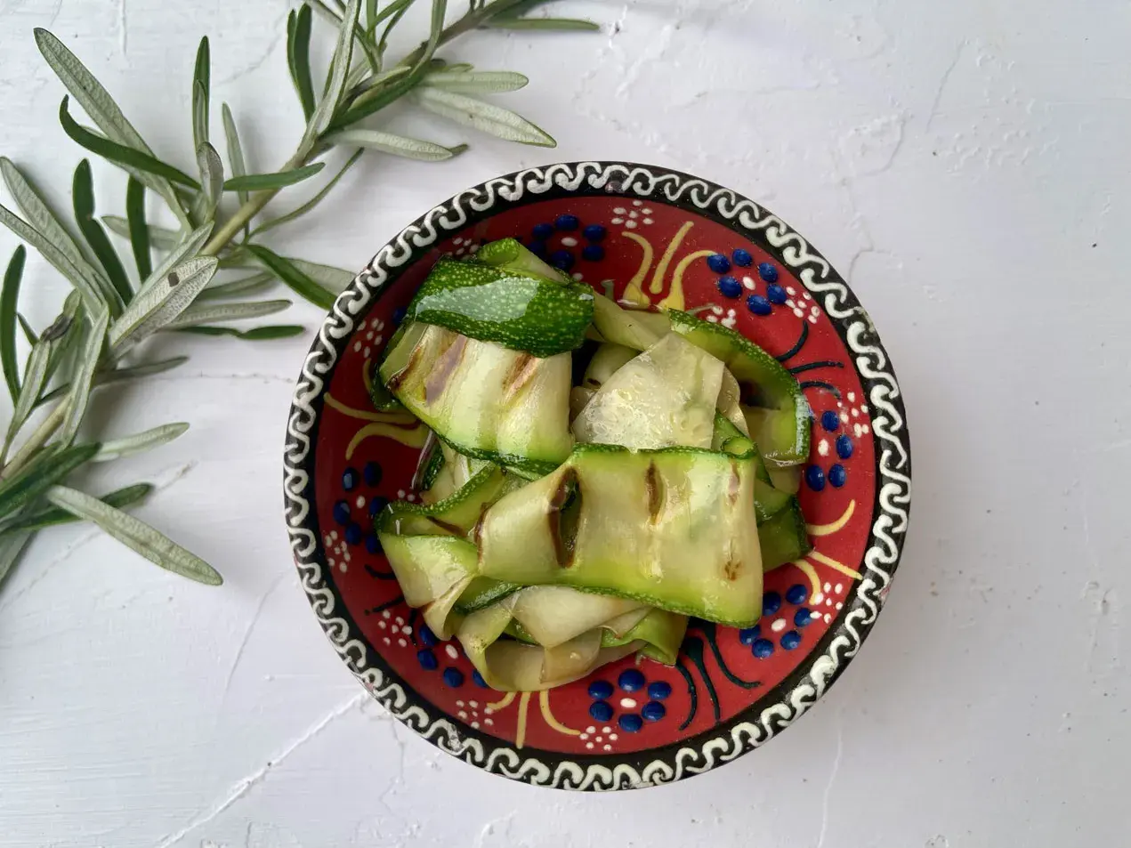 Gegrillte Zucchinibänder in einer bunten Schale, daneben Rosmarin. Perfekt, um zu sehen, was passt zu Zucchini!