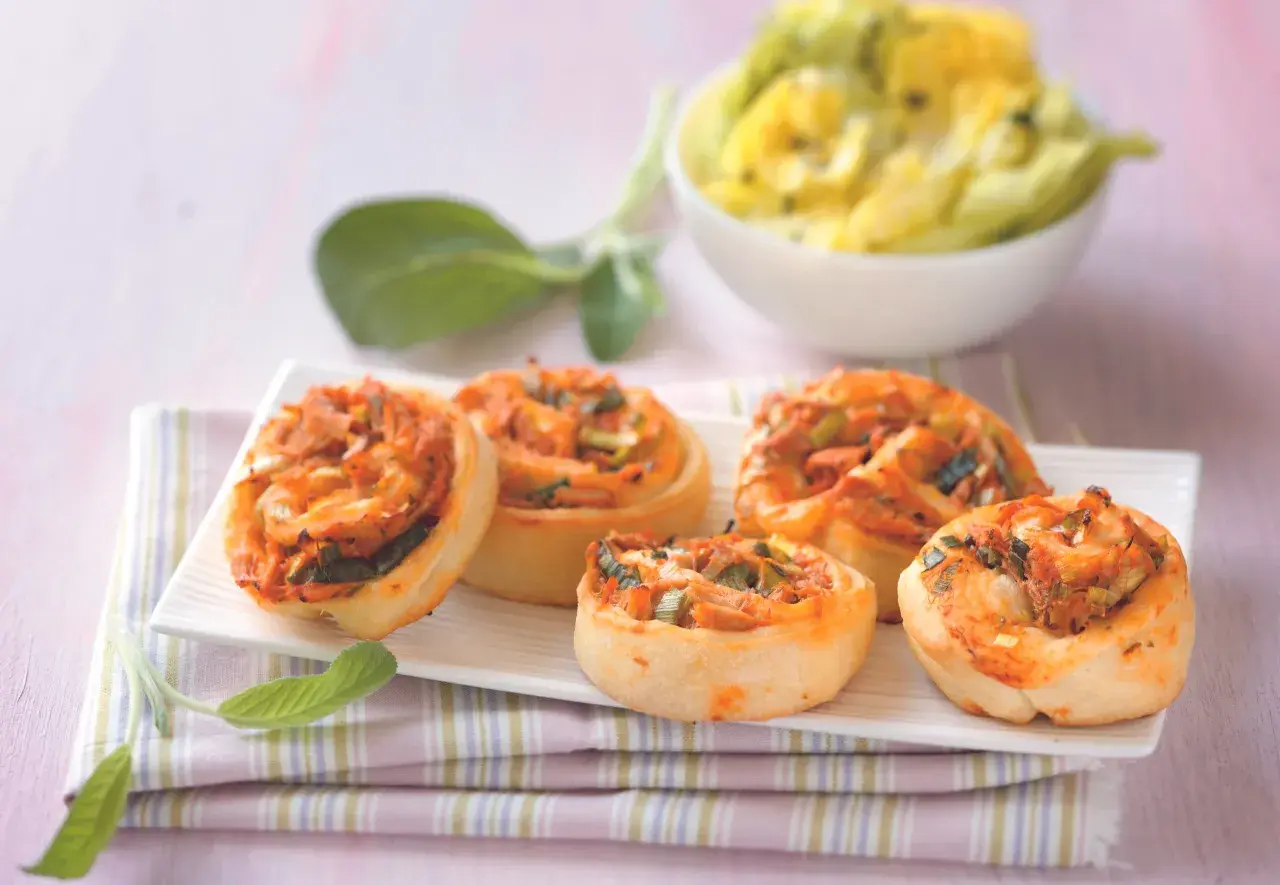 Fünf leckere Pizzaschnecken Thunfisch mit Tomaten und Lauchzwiebeln auf einem weißen Teller, daneben ein Schälchen mit gelbem Dip.