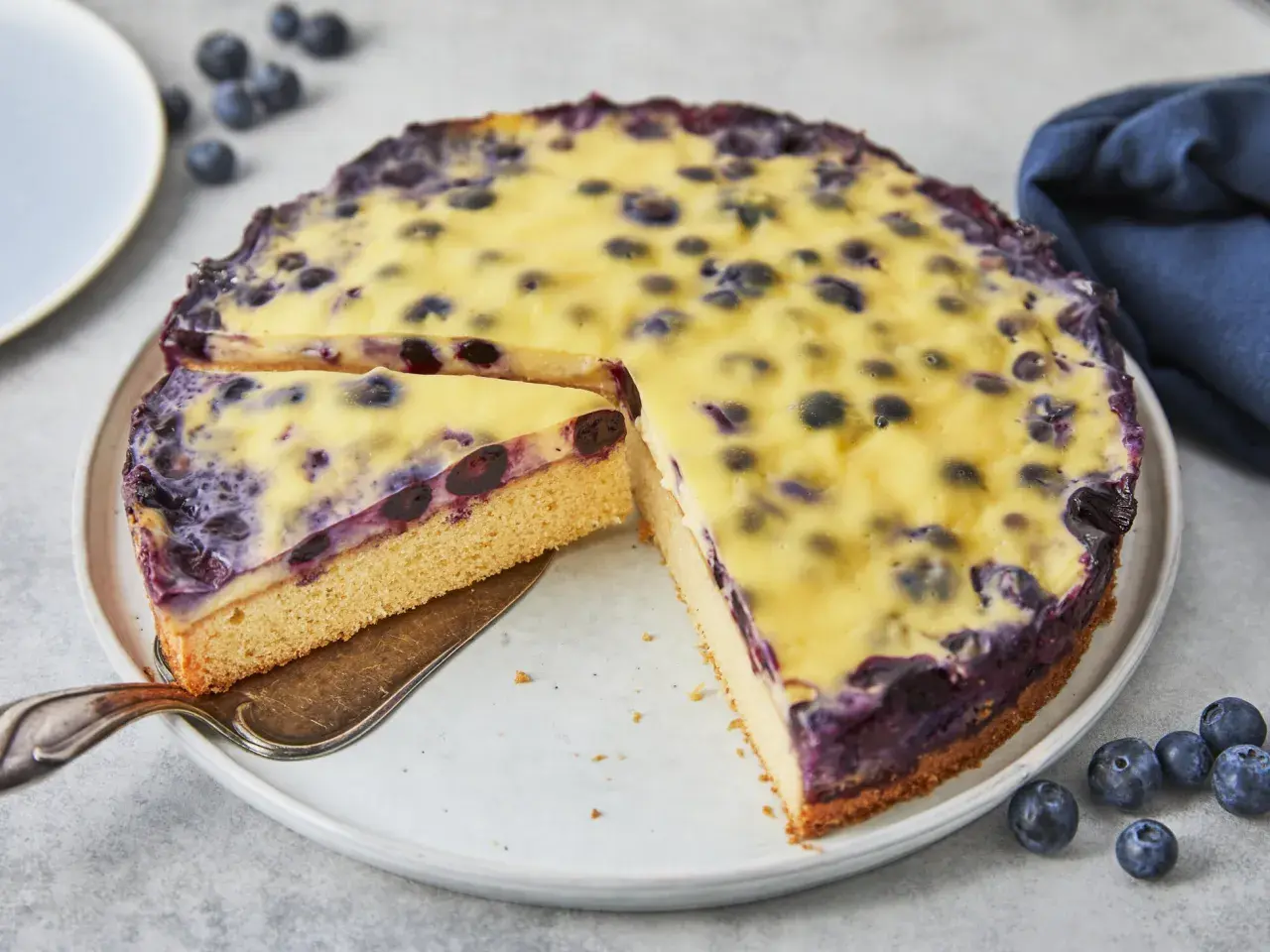 Ein saftiger Heidelbeerkuchen mit cremigem Vanillepudding, ein Stück wird gerade herausgehoben.