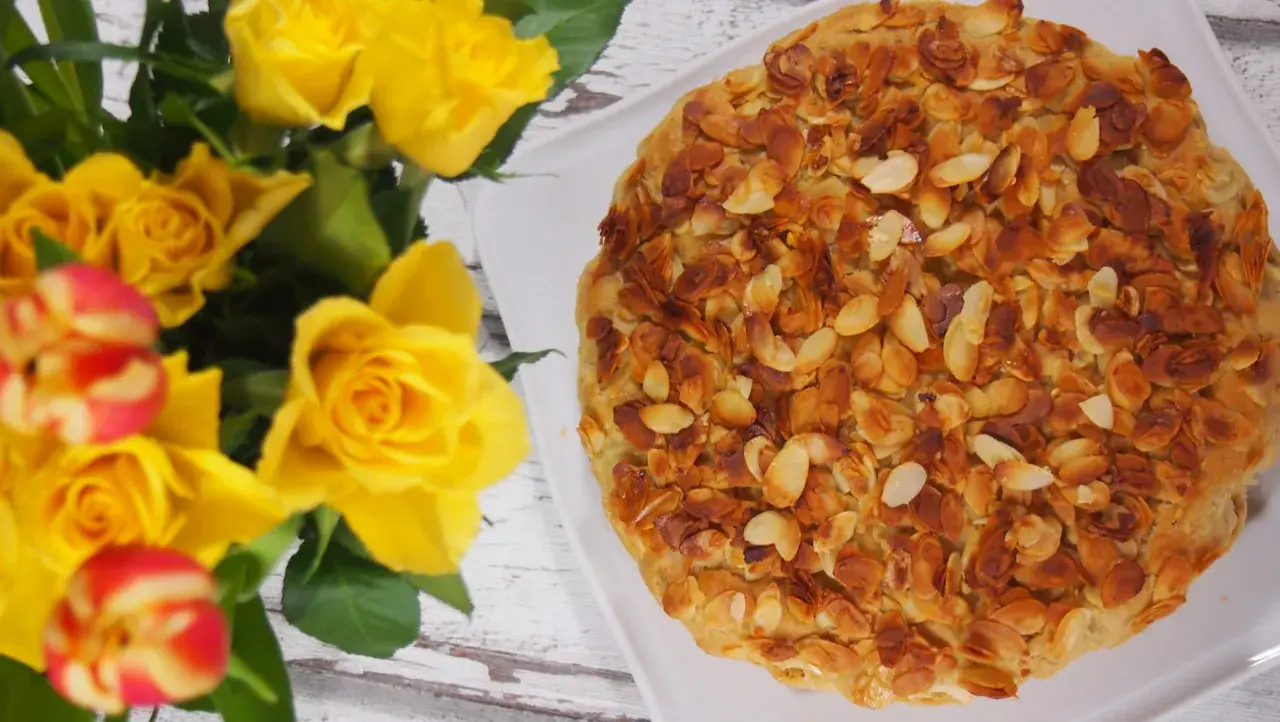 Bienenstich mandelkruste ohne backen, verziert mit gerösteten Mandelsplittern. Daneben ein Strauß gelber Rosen und Tulpen.
