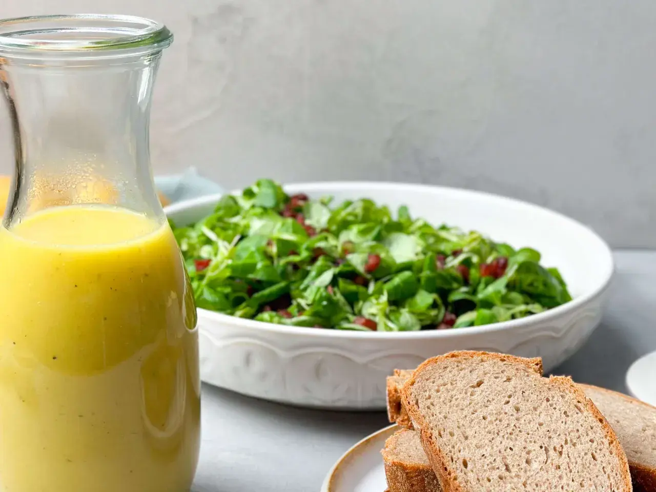 Ein Glas mit gelbem Dressing steht neben einem Salat und Brot. Perfekt für ein Kartoffel Dressing!
