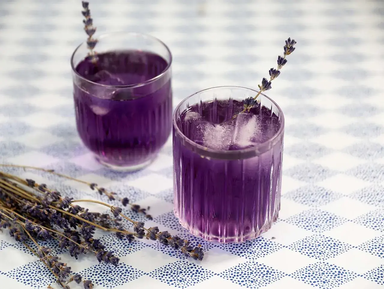 Zwei Gläser mit lila Lavendel Spritz, Eiswürfeln und Lavendelzweigen auf kariertem Tisch.